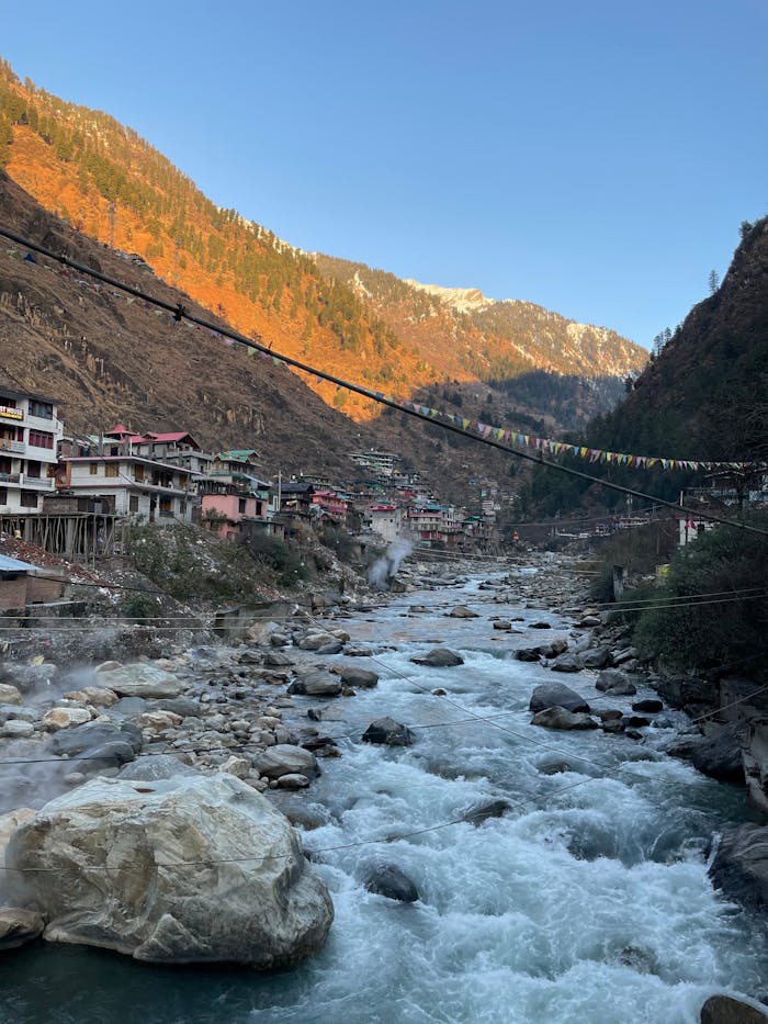 gallery-3 Serene view of a mountain village along a flowing river under a clear blue sky.